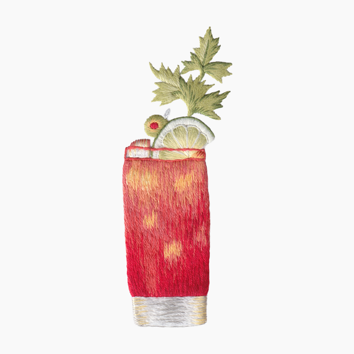 Hand embroidered cotton towel with red and orange Bloody Mary cocktail with a garnish of celery, lime and green olive on a white background